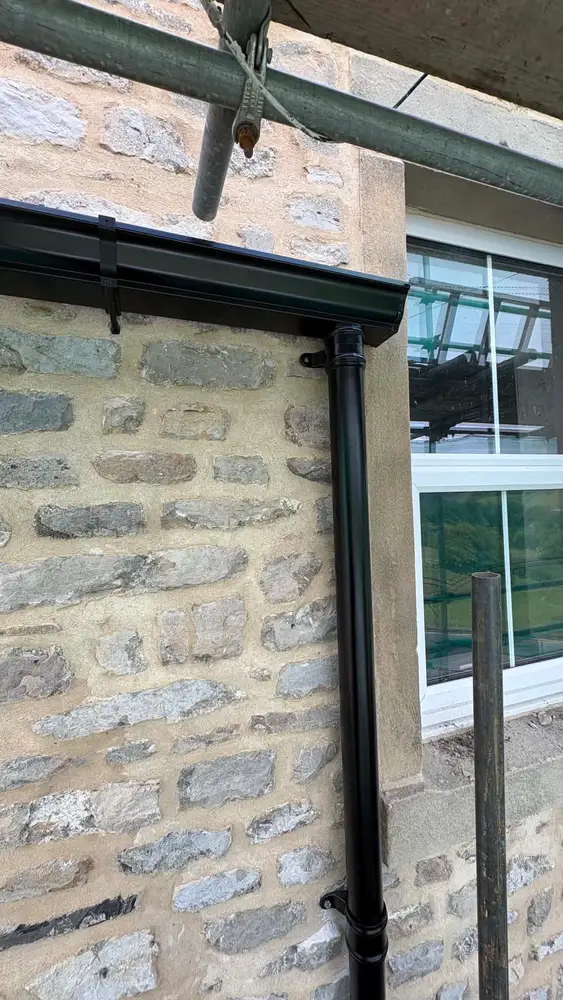 Black seamless guttering and downpipe fitted to a traditional stone wall